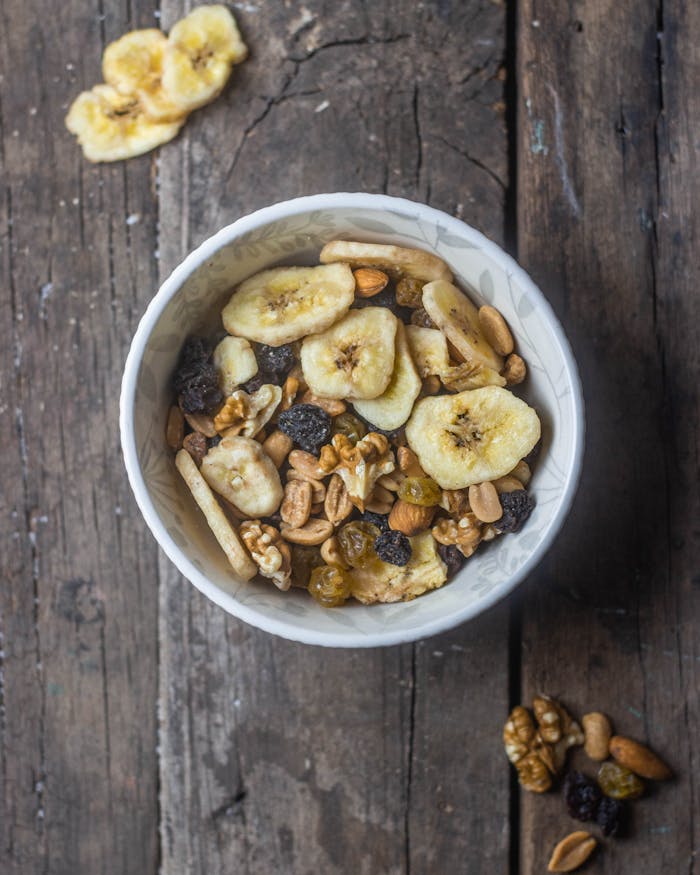 Top view of a bowl filled with mixed nuts and dried fruits including bananas, on a rustic wooden table.